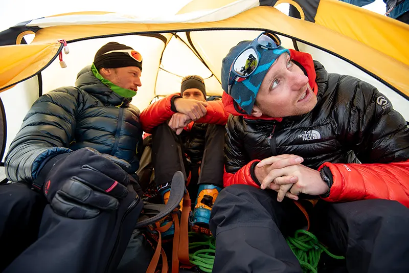 Drie mensen in winterkleding zitten dicht bij elkaar in een gele tent, met rondom hen klimtouwen en klimuitrusting. Ze lijken zich voor te bereiden op een avontuur in de buitenlucht of daar even bij uit te rusten.